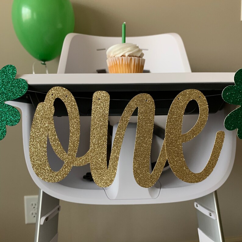 May include: Gold glitter "one" banner with green shamrock accents, perfect for a first birthday party.