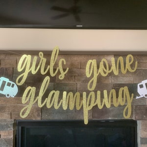 May include: A gold glitter banner with the words "girls gone glamping" in cursive script. Two camper trailer cutouts flank the text. The banner is strung across a stone fireplace.