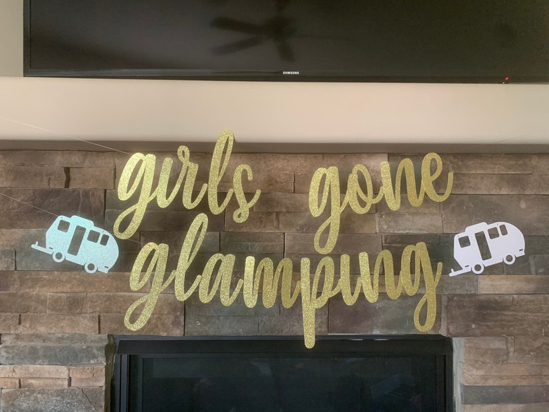 May include: A gold glitter banner with the words "girls gone glamping" in cursive script. Two camper trailer cutouts flank the text. The banner is strung across a stone fireplace.