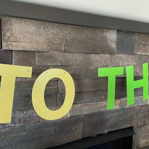 May include: A banner with the words "TO THE" in large, glittery letters. The letters "TO" are gold, and "THE" is bright green. The banner is strung against a stone wall.