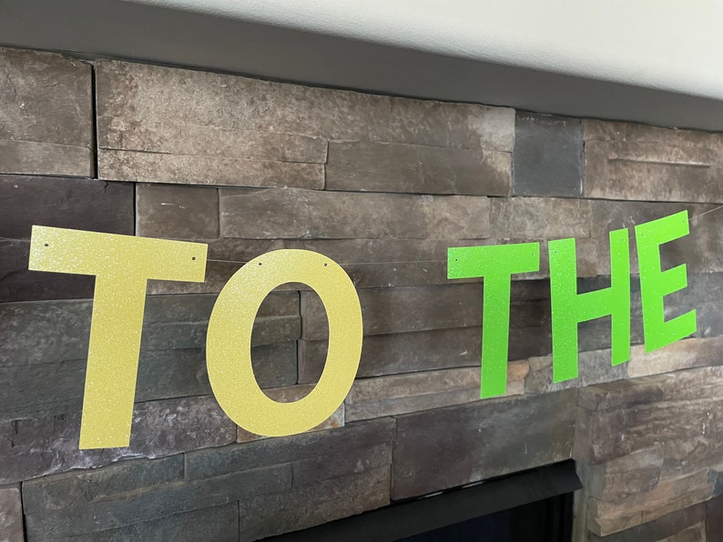 May include: A banner with the words "TO THE" in large, glittery letters. The letters "TO" are gold, and "THE" is bright green. The banner is strung against a stone wall.