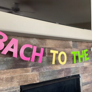 May include: A colorful banner with the text "BACH TO THE 80'S" in glittery pink, yellow, green, and blue letters. The banner is hanging in front of a stone fireplace and a black television.