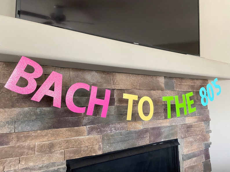 May include: A colorful banner with the text "BACH TO THE 80'S" in glittery pink, yellow, green, and blue letters. The banner is hanging in front of a stone fireplace and a black television.