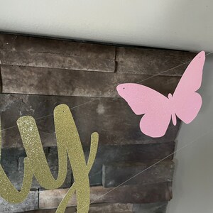 May include: A banner with gold glitter lettering and a pink glitter butterfly. The banner is strung on a clear string and is hanging in front of a stone wall. The banner is for a party.