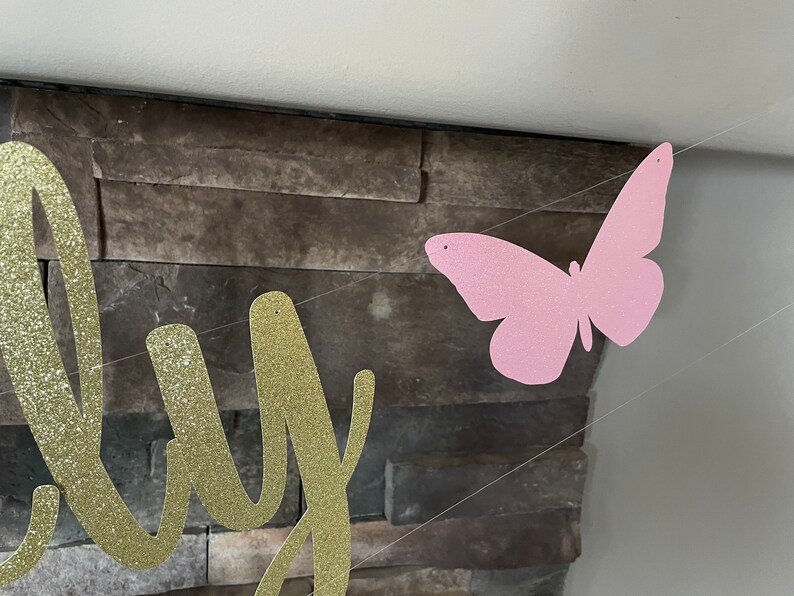 May include: A banner with gold glitter lettering and a pink glitter butterfly. The banner is strung on a clear string and is hanging in front of a stone wall. The banner is for a party.