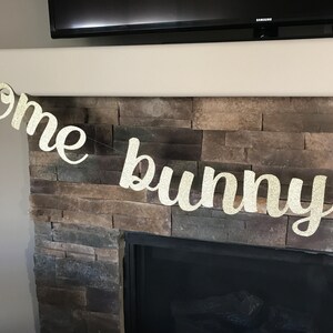 May include: A glittery gold banner that reads "Some bunny" in a cursive font. The banner is strung across a fireplace with a stone facade. The banner is likely for an Easter or baby shower celebration.