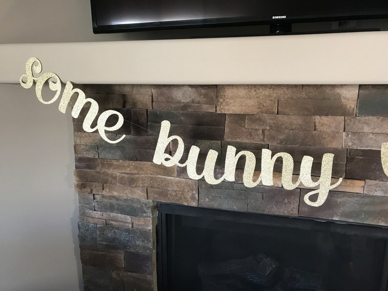 May include: A glittery gold banner that reads "Some bunny" in a cursive font. The banner is strung across a fireplace with a stone facade. The banner is likely for an Easter or baby shower celebration.