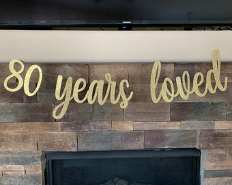 May include: A gold glitter banner that reads "80 years loved" in a cursive font. The banner is strung across a stone fireplace. The numbers and words are connected and have a sparkly finish.