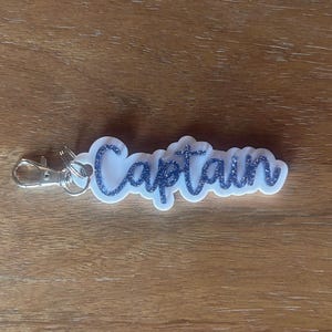 May include: A white keychain with the word "Captain" in blue glitter script. The keychain has a silver clasp for attaching to keys or bags. The design is on a white, cloud-shaped background.