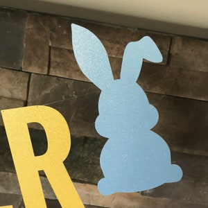 May include: A light blue glittery bunny silhouette and gold glitter letter 'R' from a banner. The bunny is on a string, suggesting a festive decoration. The background is a textured, brown stone wall.