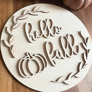 May include: A wooden round sign with the words "hello fall" in a cursive font. The sign is decorated with a pumpkin and leaves.