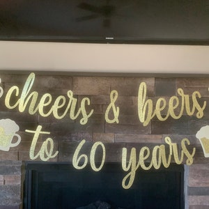 May include: A gold glitter banner with the words "cheers & beers to 60 years" in a cursive font. The banner includes cutouts of beer mugs with glitter foam. The banner is hanging in front of a stone fireplace.