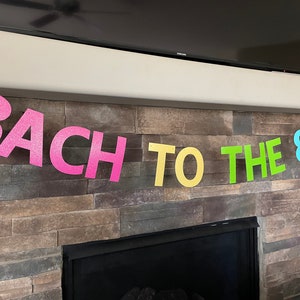 May include: A colorful banner with the text "BACH TO THE 80'S" in pink, yellow, green, and blue glitter letters. The banner is strung across a stone fireplace, perfect for a party.
