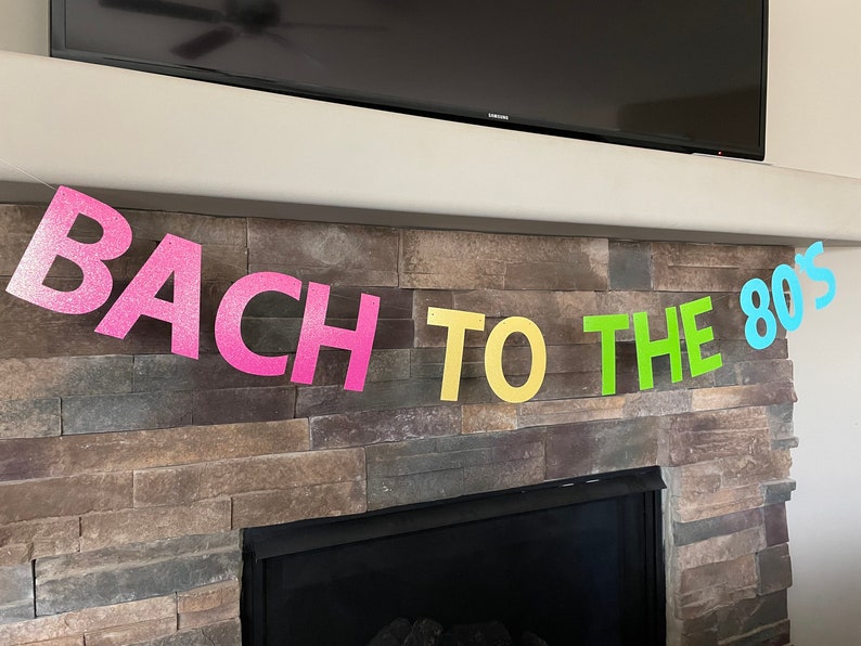 May include: A colorful banner with the text "BACH TO THE 80'S" in pink, yellow, green, and blue glitter letters. The banner is strung across a stone fireplace, perfect for a party.