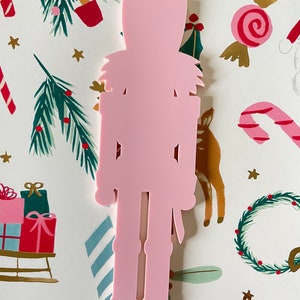 May include: A pink wooden nutcracker ornament. The nutcracker is standing upright with its arms at its sides. The nutcracker has a hat, a mustache, and a sword.