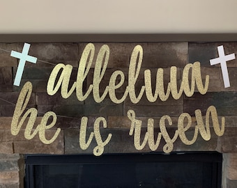 May include: A gold glitter banner with the words "alleluia he is risen" in cursive script, strung between two white crosses. The banner is displayed against a stone fireplace.
