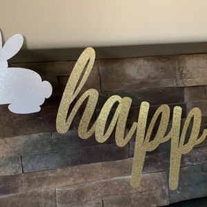 May include: A glittery gold "happy" banner and a white glitter bunny silhouette are strung together. The banner is in a cursive font. The background is a stone wall, creating a festive Easter or spring decoration.