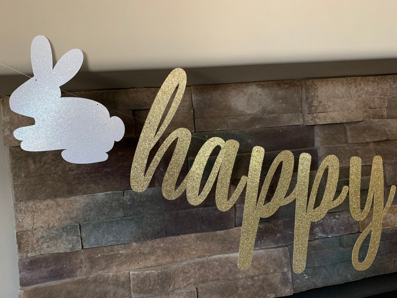May include: A glittery gold "happy" banner and a white glitter bunny silhouette are strung together. The banner is in a cursive font. The background is a stone wall, creating a festive Easter or spring decoration.
