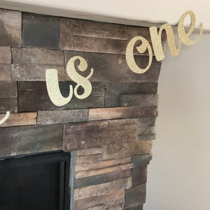May include: A gold glitter banner that reads "us one" is displayed against a stone fireplace. The banner is made of a sparkly material and is in a cursive font. The banner is likely for a first birthday celebration.