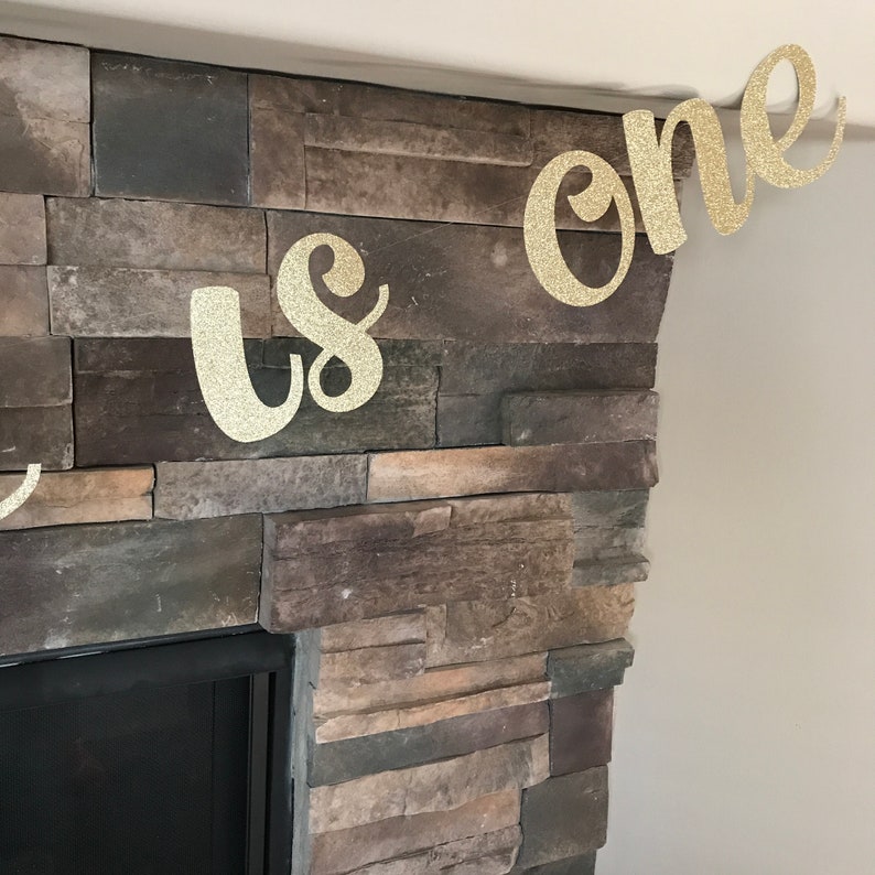 May include: A gold glitter banner that reads "us one" is displayed against a stone fireplace. The banner is made of a sparkly material and is in a cursive font. The banner is likely for a first birthday celebration.