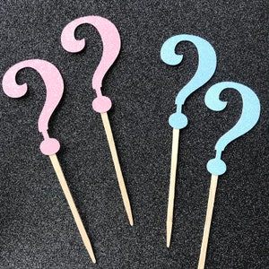 May include: Four question mark shaped party decorations, two are pink and two are blue. The decorations are made of glitter cardstock and have wooden sticks attached to the back.