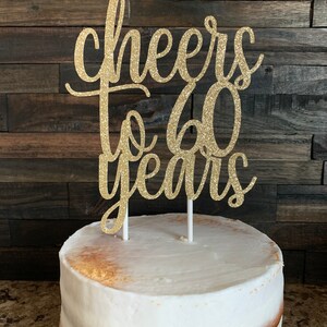 Może przedstawiać: Złoty brokatowy topper na ciasto z napisem "cheers to 60 years" umieszczony jest na wierzchu białego lukrowanego ciasta. Ciasto ma p&oacute;łnagą konstrukcję z widocznymi warstwami i stoi na czarnej paterze. Tło to ciemny panel drewniany.