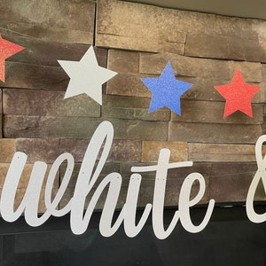 May include: A patriotic banner with red, white, and blue glitter stars strung together. The banner spells out "white &" in a cursive font. The banner is displayed against a stone wall.