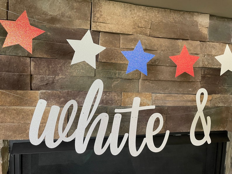May include: A patriotic banner with red, white, and blue glitter stars strung together. The banner spells out "white &" in a cursive font. The banner is displayed against a stone wall.
