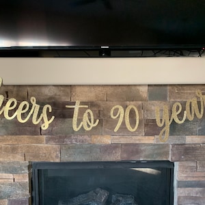 May include: A gold glitter banner that reads "cheers to 90 years" is strung across a stone fireplace. The banner is in a cursive font and is centered above the fireplace. A black television is mounted above the fireplace.