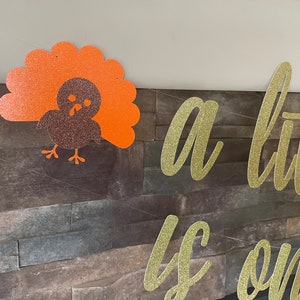May include: Thanksgiving banner with a glittery turkey decoration and the phrase "a little is on the way" in gold glitter lettering. The turkey is brown and orange, and the banner is strung on a thin white string.