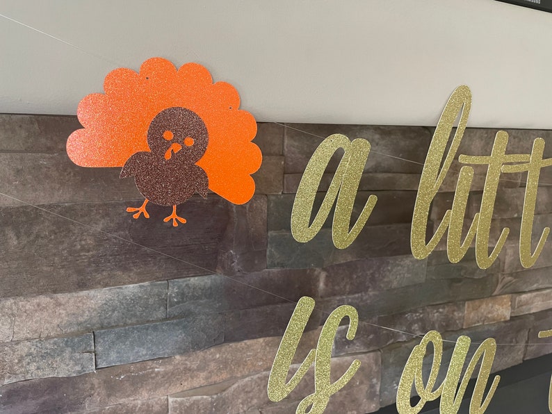 May include: Thanksgiving banner with a glittery turkey decoration and the phrase "a little is on the way" in gold glitter lettering. The turkey is brown and orange, and the banner is strung on a thin white string.