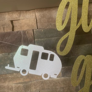 May include: A glittery white camper trailer banner with a gold glitter cursive "go" banner. The camper has a door, windows, and wheels. The banners are strung on a thin white string, set against a textured stone wall.