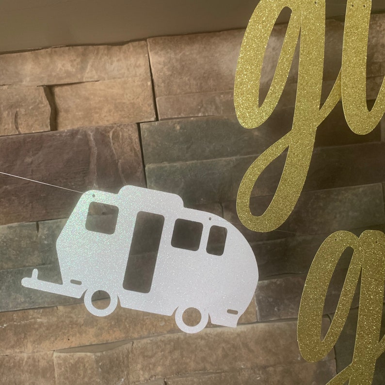 May include: A glittery white camper trailer banner with a gold glitter cursive "go" banner. The camper has a door, windows, and wheels. The banners are strung on a thin white string, set against a textured stone wall.