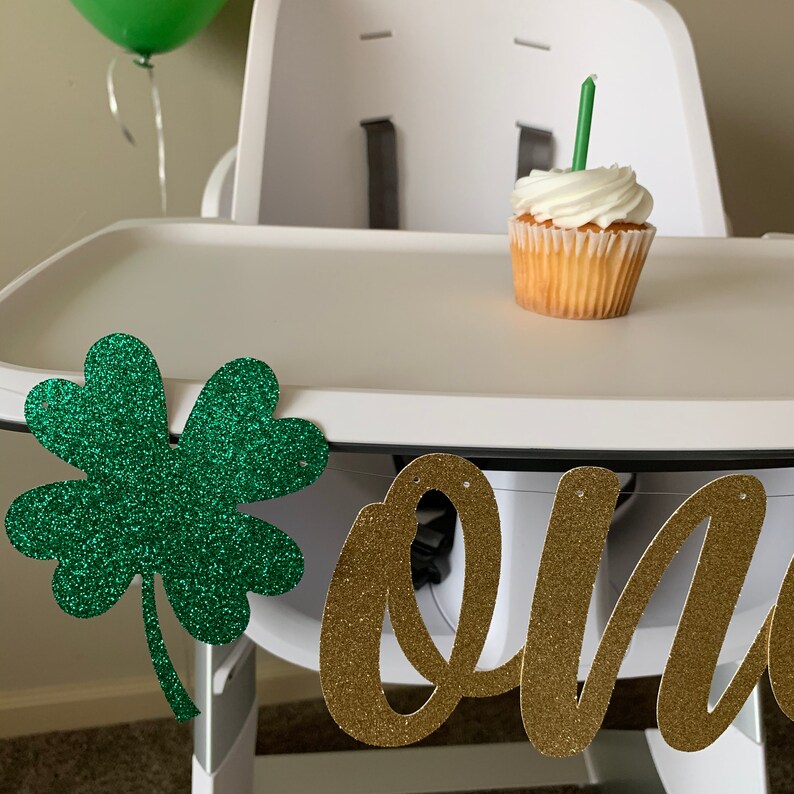 May include: A white high chair with a green shamrock and gold glitter "one" banner. A cupcake with green frosting and a single candle sits on the tray.