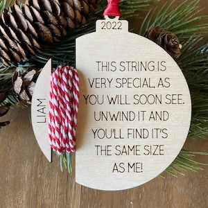May include: Wooden Christmas ornament with a red and white string attached. The ornament is shaped like a circle with a cut-out section. The text on the ornament reads: "This string is very special, as you will soon see. Unwind it and you'll find it's the same size as me!" The year 2022 is also printed on the ornament.