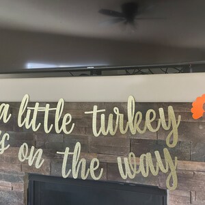 May include: A gold glitter banner with the phrase "a little turkey is on the way" in cursive script, flanked by orange turkey cutouts. The banner is displayed above a fireplace.