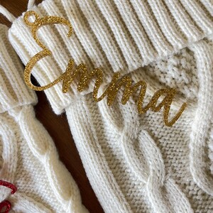 May include: White knitted stocking with a gold glitter name tag that reads "Emma".