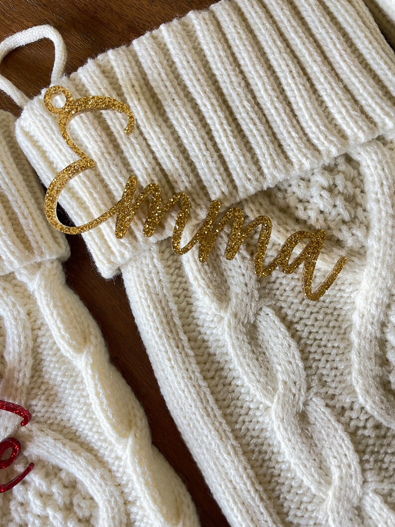 May include: White knitted stocking with a gold glitter name tag that reads "Emma".