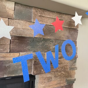 May include: A festive banner with glittery star cutouts in white, red, and blue, spelling out the word "TWO". The banner is strung across a stone fireplace, perfect for a second birthday celebration.