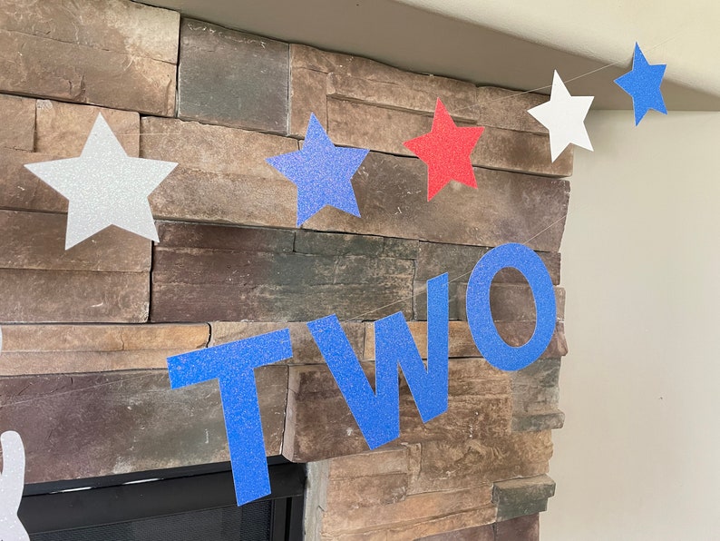 May include: A festive banner with glittery star cutouts in white, red, and blue, spelling out the word "TWO". The banner is strung across a stone fireplace, perfect for a second birthday celebration.