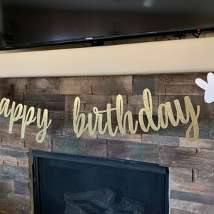 May include: A gold glitter "happy birthday" banner with white bunny cutouts on each end. The banner is strung across a fireplace with a stone facade. The text is in a cursive font.