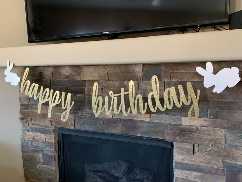 May include: A gold glitter "happy birthday" banner with white bunny cutouts on each end. The banner is strung across a fireplace with a stone facade. The text is in a cursive font.