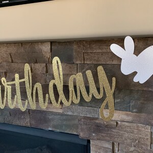 May include: A gold glitter "birthday" banner and a white glitter bunny decoration hang against a stone wall. The banner is in a cursive font. The bunny is a silhouette. The decorations are likely for a birthday party.