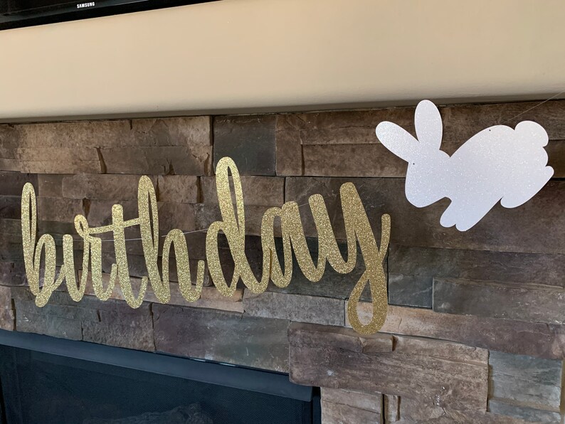 May include: A gold glitter "birthday" banner and a white glitter bunny decoration hang against a stone wall. The banner is in a cursive font. The bunny is a silhouette. The decorations are likely for a birthday party.