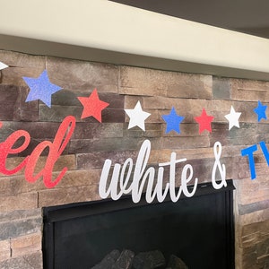 May include: A patriotic banner with red, white, and blue glitter stars and the words "red white & TWO". The banner is strung across a fireplace with a stone facade, perfect for a second birthday celebration.