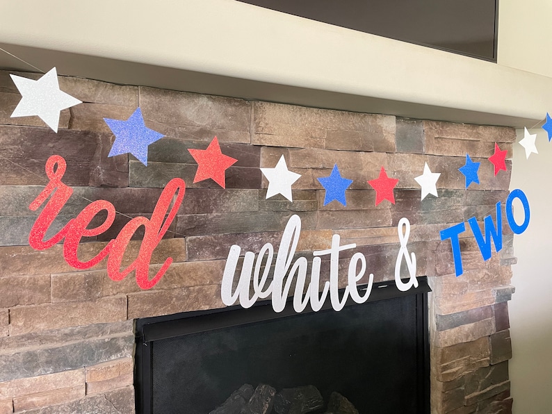 May include: A patriotic banner with red, white, and blue glitter stars and the words "red white & TWO". The banner is strung across a fireplace with a stone facade, perfect for a second birthday celebration.