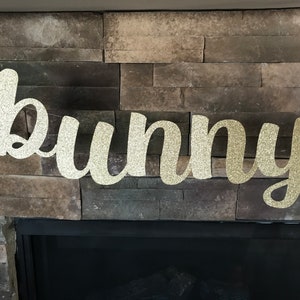 May include: A glittery gold "bunny" banner, perfect for Easter decorations. The word is written in a cursive font and strung on a thin, nearly invisible string. The banner is displayed against a backdrop of a stone fireplace.
