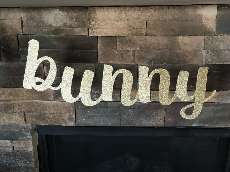 May include: A glittery gold "bunny" banner, perfect for Easter decorations. The word is written in a cursive font and strung on a thin, nearly invisible string. The banner is displayed against a backdrop of a stone fireplace.