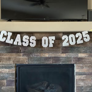 May include: A silver glitter banner with the text "Class of 2025" hanging above a fireplace. The banner is decorated with two gold glitter graduation caps.