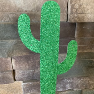 K&ouml;nnte beinhalten: Eine glitzernde, gr&uuml;ne Kaktus-Dekoration. Der Kaktus hat eine strukturierte, funkelnde Oberfl&auml;che und ist vor einem Hintergrund aus gestapelten Steinen aufgeh&auml;ngt. Das Design zeigt die klassische Form eines Saguaro-Kaktus.